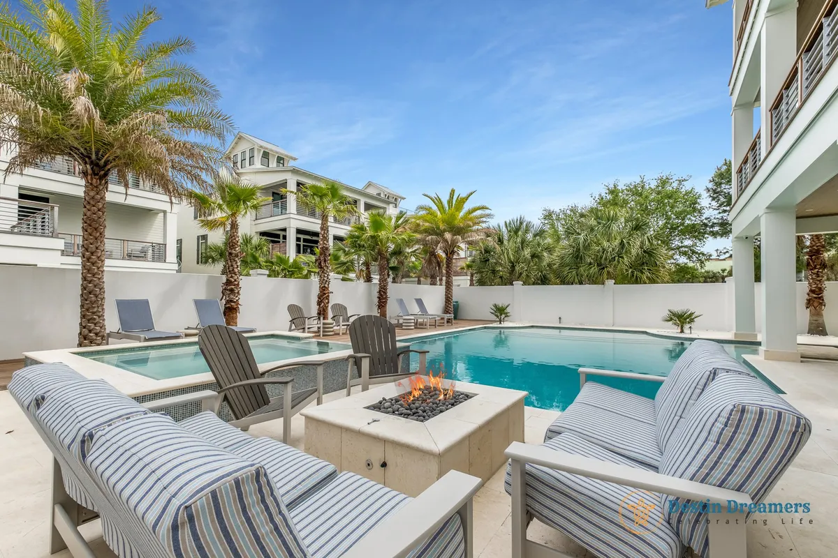 A luxurious backyard space with a sparkling pool, a cozy fire pit surrounded by outdoor seating, and lush palm trees swaying in the evening light. The ambiance is relaxed and inviting, encouraging both relaxation and socialization in a resort-style environment.