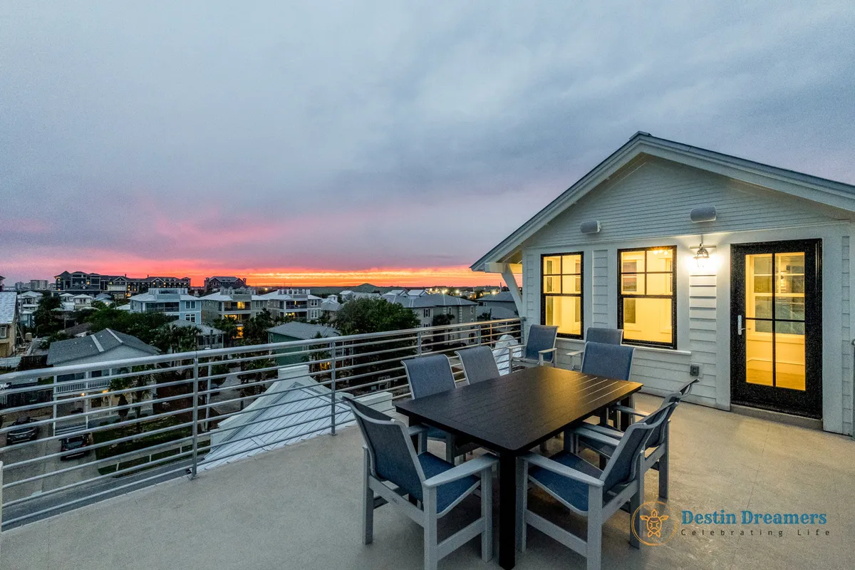 An elegant outdoor dining table set beneath an open sky, with the sun setting over the ocean in the distance. Modern architectural elements frame the view, and the scene radiates a sense of tranquil togetherness—ideal for gathering with friends or family after a day of complimentary activities.