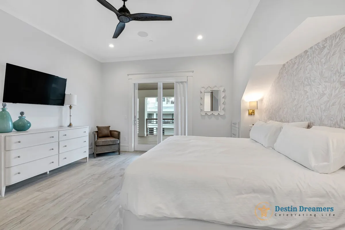 A serene king suite featuring a king-sized bed, decorative wall panel, television, lounge chair, and sliding glass doors that open to a balcony. The ambiance is tranquil, offering a private retreat within the home.