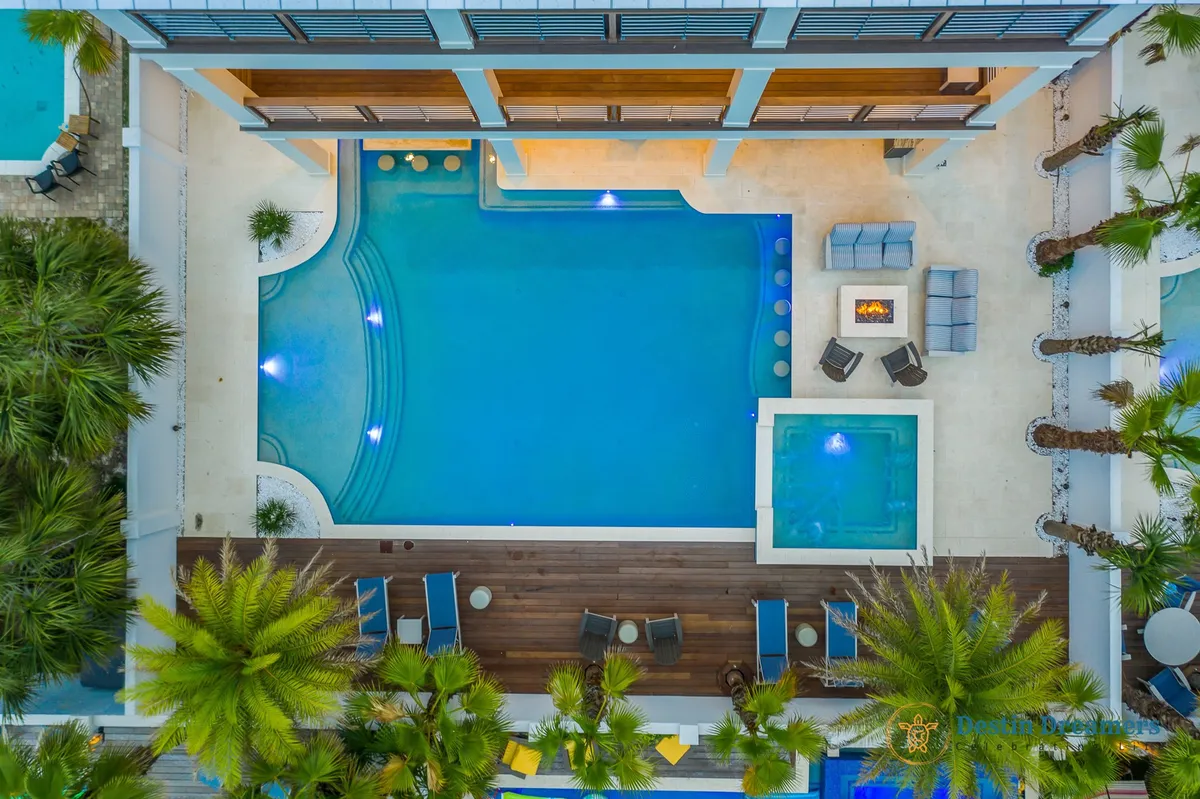 An inviting exterior space featuring a swimming pool, a hot tub, a cozy fire pit, and lush palm landscaping. The area is designed for socializing, relaxation, and evening gatherings, with atmospheric lighting enhancing the sense of warmth and luxury across all seasons.