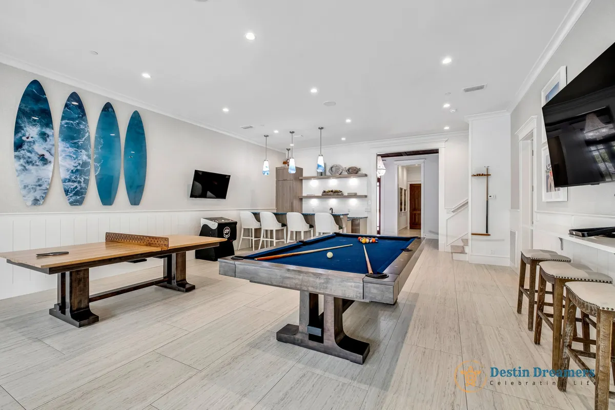 A vibrant game room featuring a sleek pool table, surfboards hung on the wall that add a playful, coastal vibe, and a built-in bar area perfect for socializing. The space is designed for fun and group interaction, with comfortable seating and a TV for additional entertainment.
