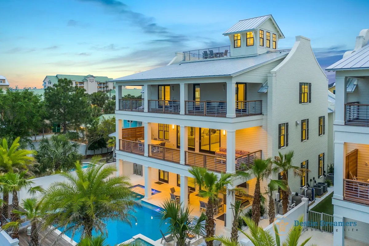 The hero image showcases a sprawling three-story coastal home surrounded by swaying palm trees, with an inviting private heated pool featuring a swim-up bar as the centerpiece. Expansive balconies overlook the sparkling pool and lush landscaping, while a rooftop crow's nest hints at breathtaking 360-degree Gulf views. The setting exudes luxury and relaxation, perfect for large groups or families eager to enjoy Destin's sun-soaked lifestyle.