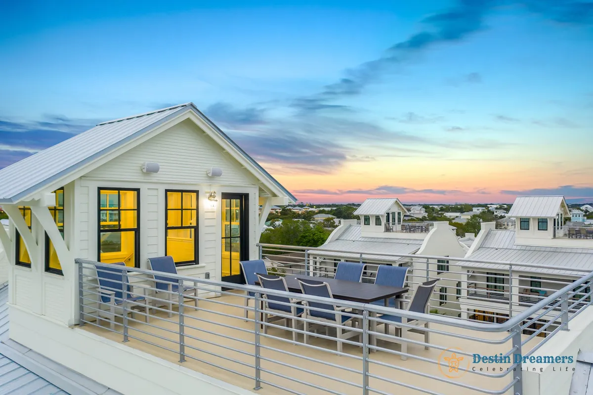 This image captures the rooftop dining table surrounded by chairs, bordered by the balcony railing, with the sunset sky painting a colorful horizon. Warm interior lighting glows through the rooftop access door, inviting in a sense of comfort and relaxation. The elevated position provides clear, panoramic views of the Gulf and cityscape.
