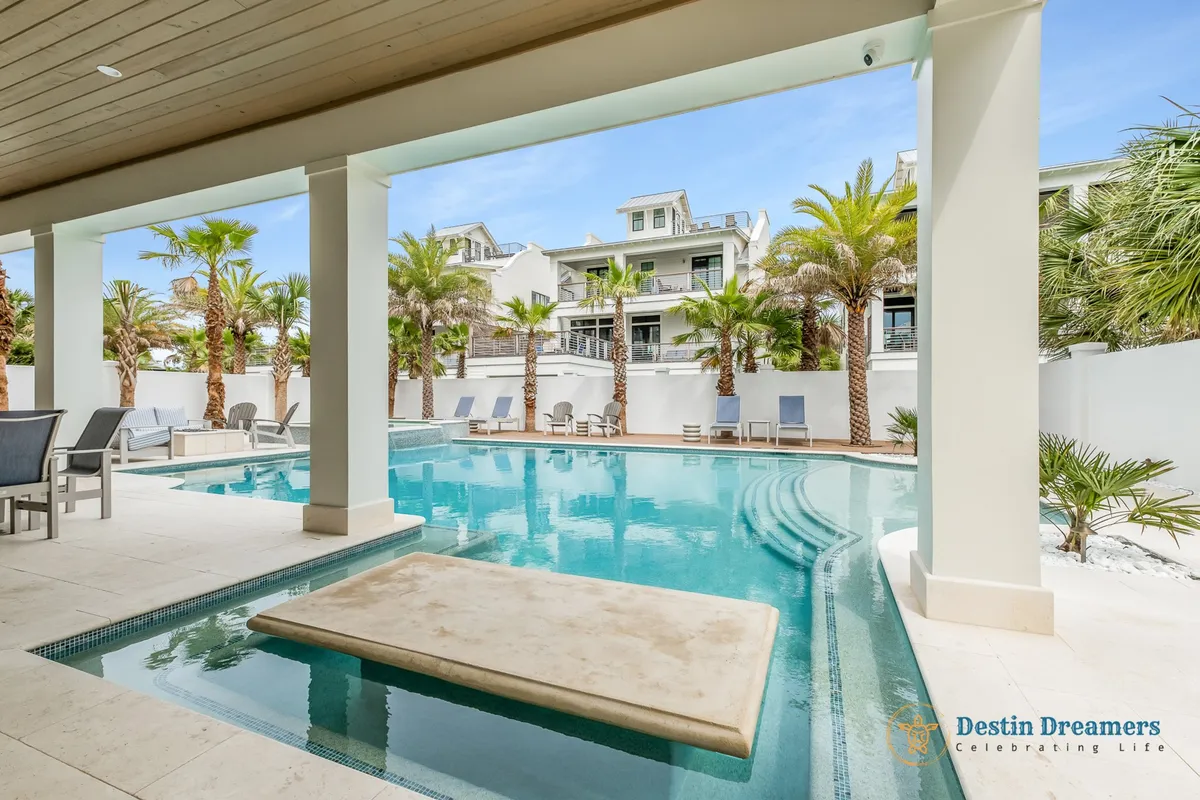 A vibrant outdoor scene featuring a private heated pool with a swim-up bar, surrounded by palm trees and a modern white multi-story vacation home with balconies. Lounge chairs and cozy seating areas create inviting spaces for sunbathing, socializing, or enjoying poolside drinks. This setting is designed for both relaxation and lively gatherings.