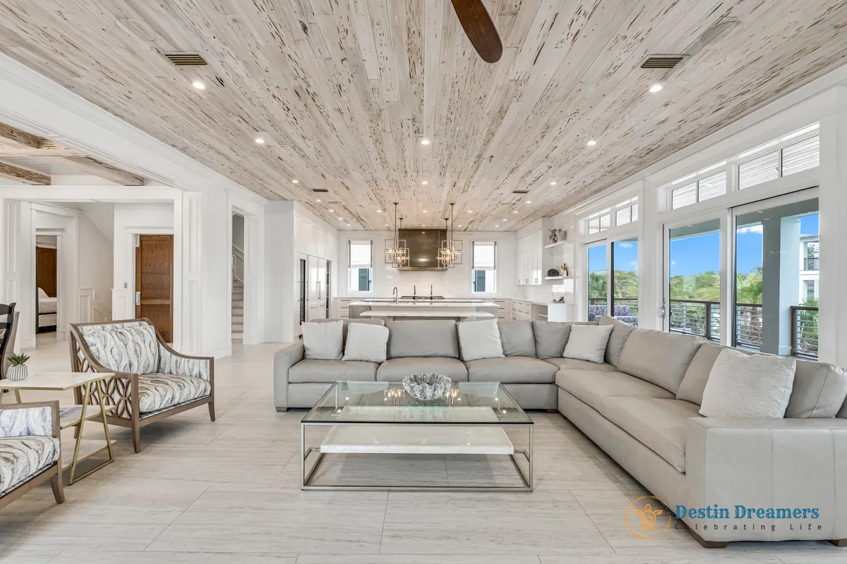 An elegant, open-concept living space with a large light gray sectional sofa, glass coffee table, open kitchen with double islands and pendant lighting, and floor-to-ceiling windows offering views and access to the balcony.