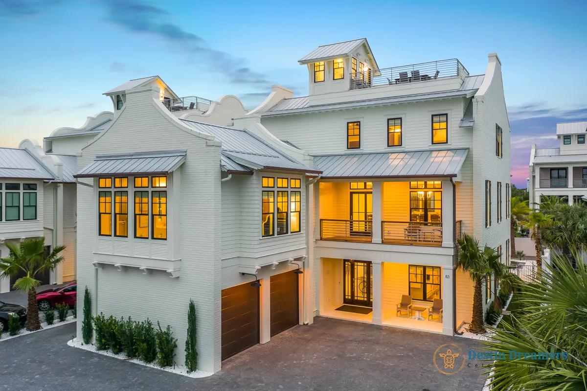 This image displays a large, multi-story white coastal home with a distinctive rooftop crow's nest. The house is surrounded by lush palm trees and coastal landscaping, with balconies and warm glowing windows hinting at comfortable interior spaces. The crow's nest atop the home is visible, suggesting the promise of panoramic Gulf and sunset views.