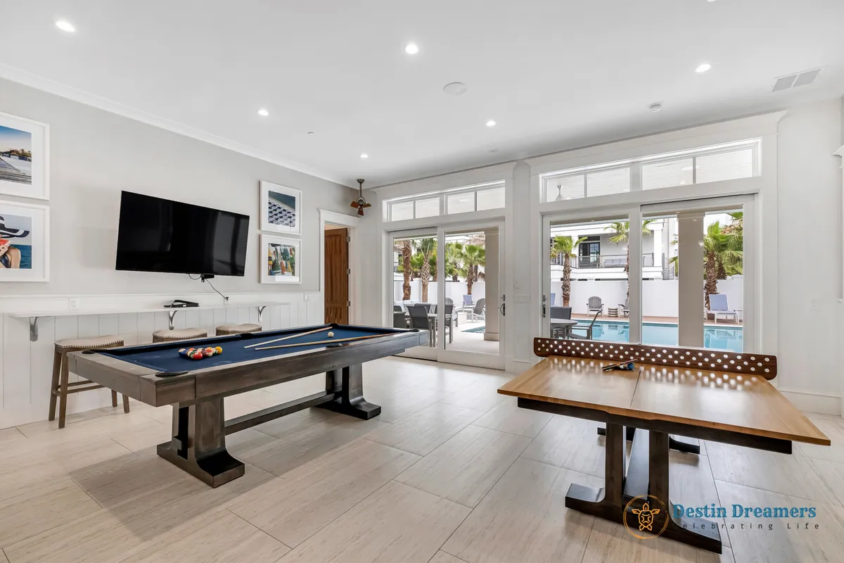 The image captures a lively game room featuring a pool table and ping pong table, with a large flat-screen TV on the wall and glass doors that open directly to the pool area. The space is bright, energetic, and designed for group entertainment and friendly competition.