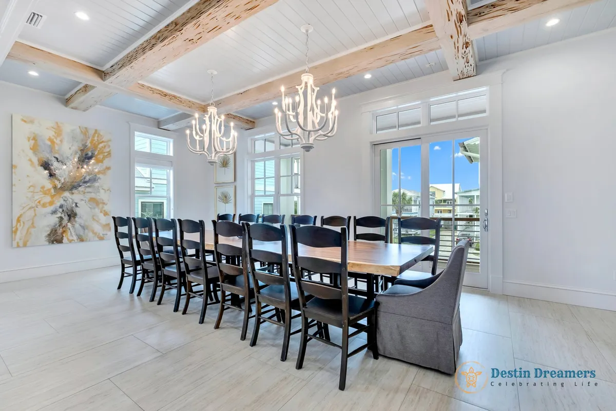 A spacious dining area featuring a long wooden table with seating for a large group, two elegant white chandeliers, abstract gold and black wall art, and multiple windows and glass doors letting in abundant natural light, with balcony access visible.