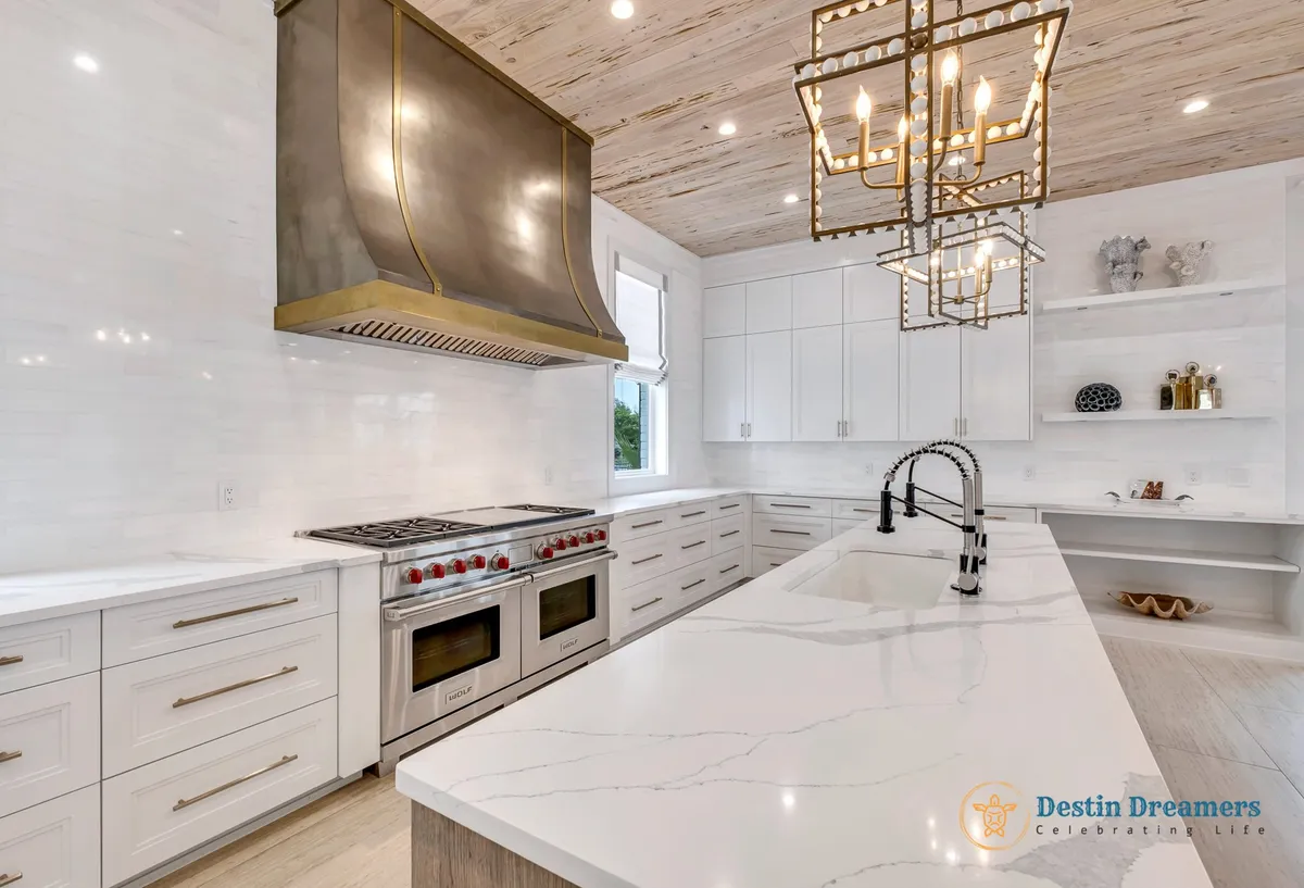 A bright, high-end kitchen with a large white marble island featuring dual faucets, professional-grade stainless steel double oven and stove with a brass hood, modern white cabinetry, and decorative hanging light fixtures. The open layout and sophisticated ambiance make it ideal for group cooking and culinary gatherings.