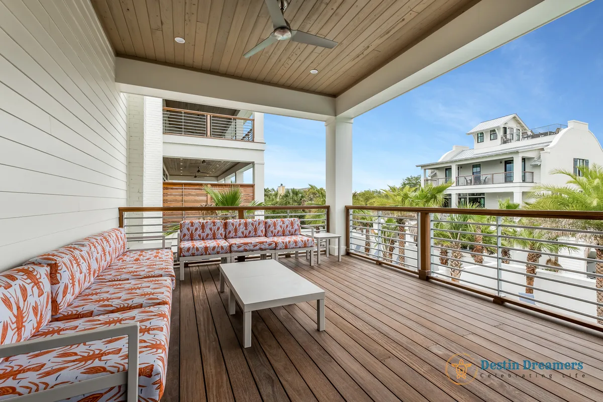 A covered balcony seating area with patterned cushions and wooden deck flooring, shaded by a ceiling fan and recessed lighting. The view includes neighboring houses and tall palm trees, evoking a peaceful, residential neighborhood vibe.