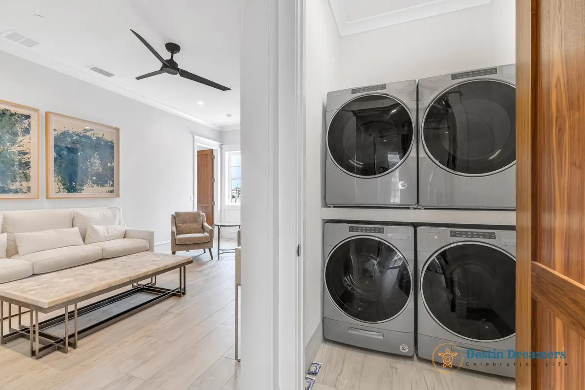 A modern laundry area with stacked washer and dryer units is conveniently located next to a comfortable living space with a beige sofa, armchair, and abstract blue artwork. The design blends functionality with comfort, supporting extended stays and family needs.