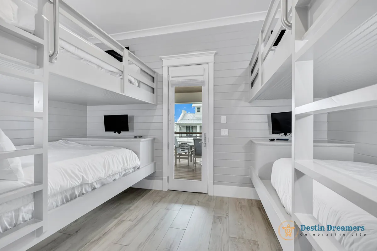 A spacious bunk room featuring two sets of white bunk beds, a glass door leading to a balcony with outdoor seating, and small flat-screen TVs on white dressers beneath the upper bunks. The room is calm, functional, and perfect for kids or groups.