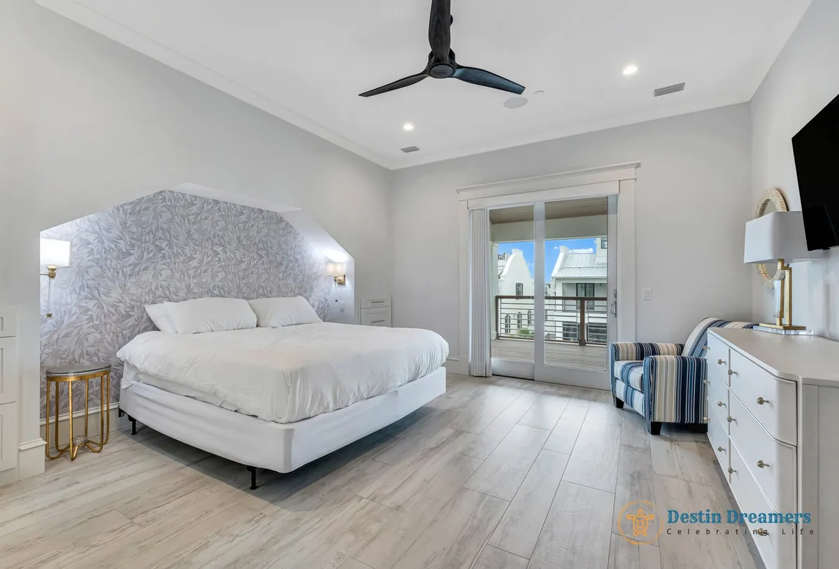 A large king-size bed with crisp white linens is set against an accent wall with a subtle leaf pattern, creating a tranquil and modern retreat. A striped armchair sits near a dresser, and a sliding glass door leads out to a private balcony, offering fresh air and views. The space feels bright, airy, and designed for ultimate relaxation.