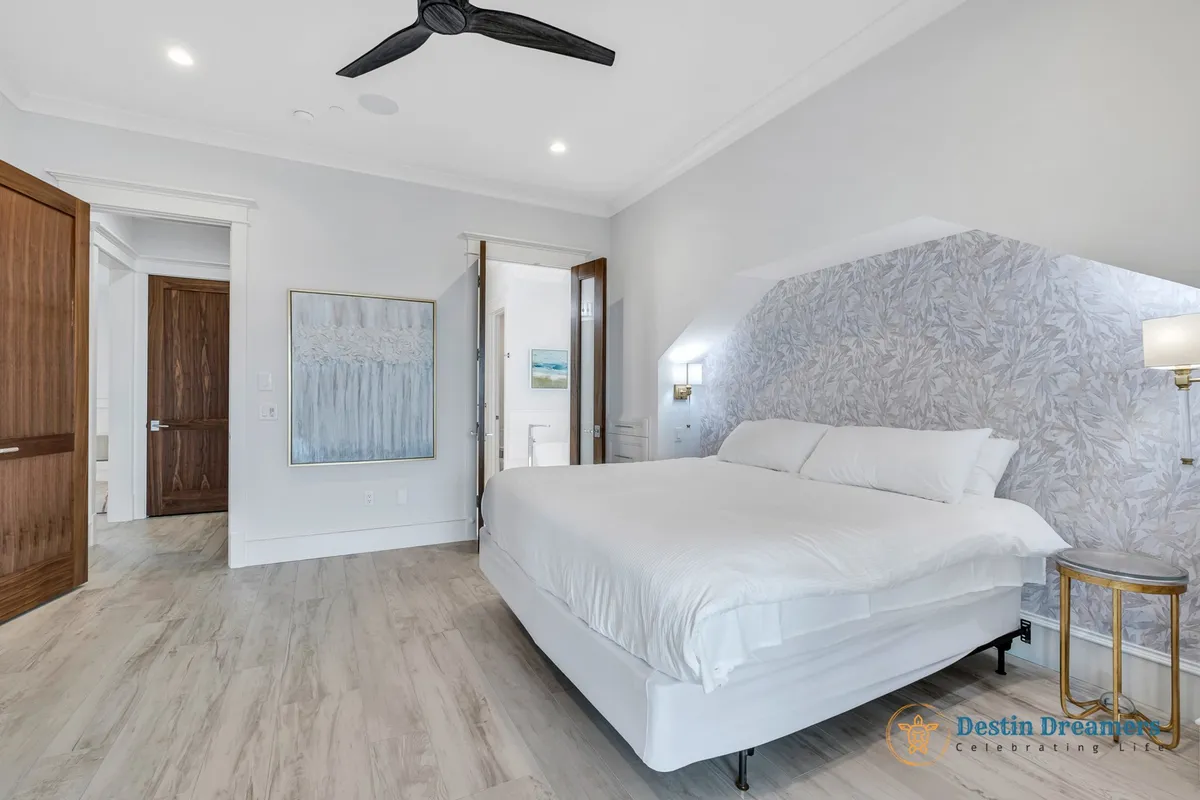 A serene king suite featuring a large king-sized bed with white linens, an accent wall with subtle patterned wallpaper, a modern ceiling fan, and open wooden doors leading to adjacent spaces, evoking comfort and privacy.
