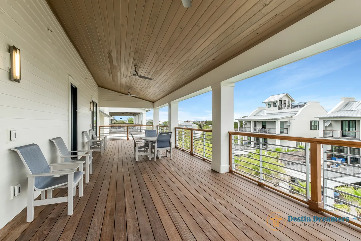 A long covered porch with wooden flooring and ceiling, furnished with outdoor seating and tables. The porch provides a view of modern coastal homes and features a stylish railing, offering a tranquil, open-air setting for leisure and socializing.