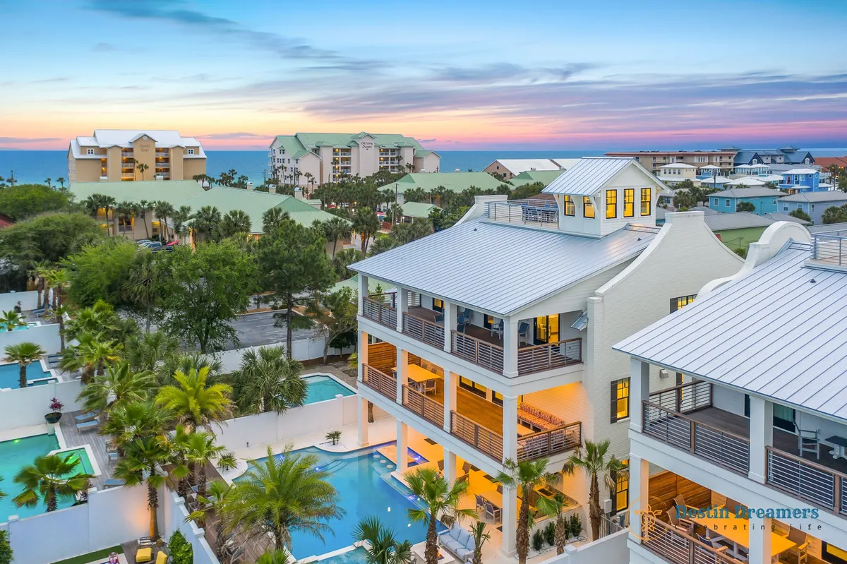 A daylight exterior shot of the multi-story vacation home, featuring wrap-around balconies, a private heated pool with a swim-up bar, and the rooftop crow's nest. Palm trees and the nearby beach are visible in the background, reinforcing the 360-degree Gulf and city views. The setup highlights spaces for both daytime poolside enjoyment and rooftop relaxation.