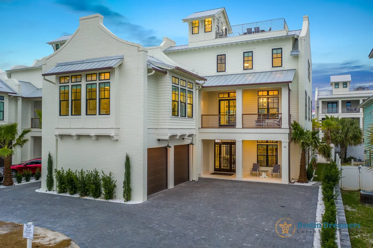 A large, white multi-story beach house with modern coastal architecture stands prominently, featuring multiple balconies with outdoor seating, a rooftop crow's nest offering panoramic views, and a wide driveway suitable for golf carts or bikes. The home is surrounded by palm trees and lush landscaping, evoking a quintessential Florida coastal vibe.