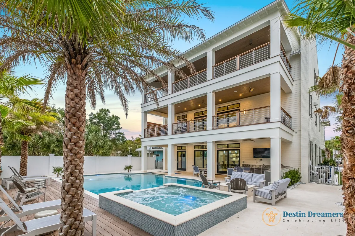 An exterior view of a private heated pool surrounded by an outdoor seating area with sofas and chairs. The modern, three-level home features multiple balconies and is framed by palm trees, creating a tropical, upscale atmosphere for outdoor activities.