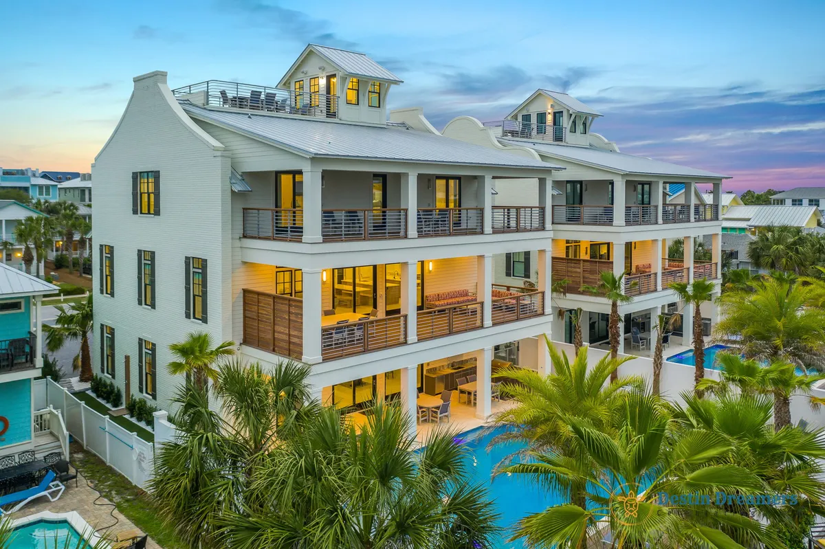 A stunning exterior view of a multi-story luxury beach house complete with a distinctive rooftop crow's nest, a large private pool with a curved design, multiple spacious balconies furnished for relaxation, and lush tropical palm trees surrounding the property. The setting exudes an upscale vacation vibe, perfect for groups and families seeking both relaxation and adventure.