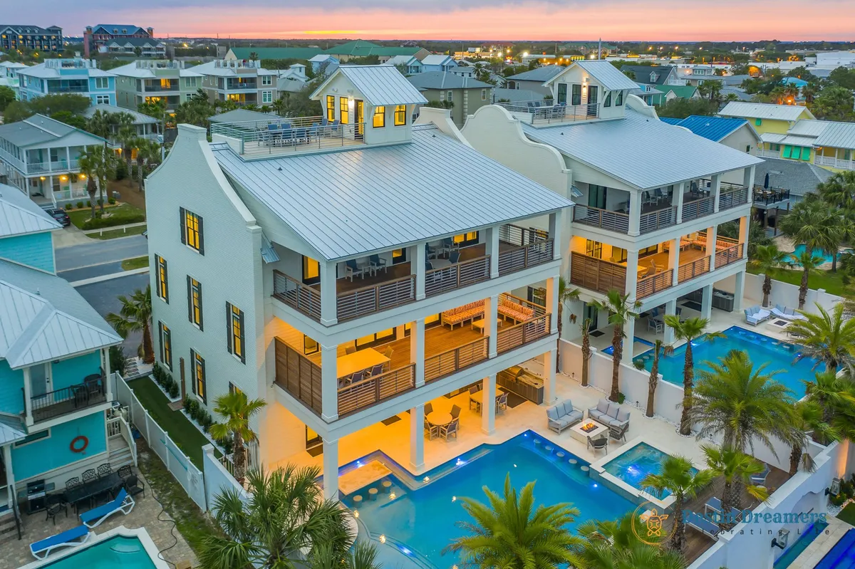 A luxurious exterior nighttime view featuring a private heated pool with a swim-up bar, multi-level balconies furnished for outdoor lounging, and a rooftop crow's nest. The home is well-lit, creating an inviting atmosphere for sunset and evening gatherings. The crow's nest and balconies indicate spaces for enjoying panoramic views, group activities, and relaxation.