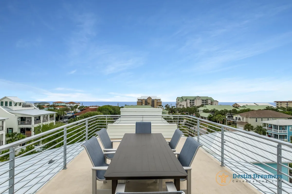 A rooftop deck with an outdoor dining table and six chairs, surrounded by a railing that frames distant ocean views and neighborhood rooftops. The open sky creates a peaceful, panoramic setting ideal for sunset meals and group gatherings.