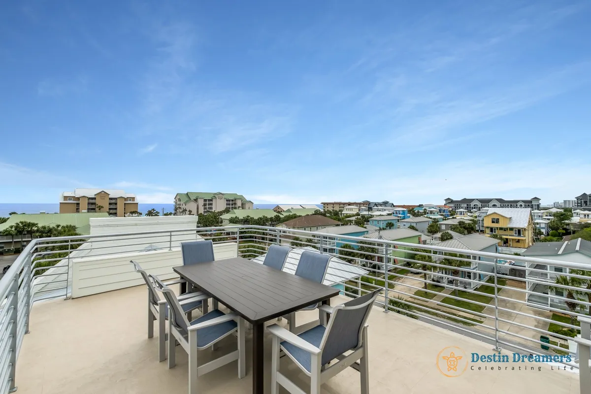 A rooftop terrace with a modern stainless steel railing and an outdoor dining table surrounded by six chairs, offering 360-degree panoramic views of the neighborhood and distant water. The clear blue sky enhances the open, airy feel, making this an ideal setting for al fresco dining or sunset gatherings.