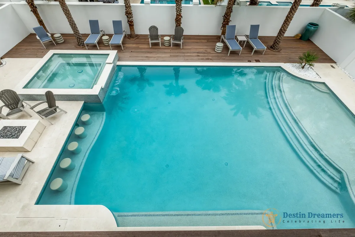 A private outdoor oasis featuring a heated pool with swim-up bar stools, an adjacent hot tub, a cozy firepit area with chairs, deck chairs for sunbathing, and palm trees providing a lush tropical backdrop. The inviting setup highlights spaces for relaxation and socializing after a day at the beach.